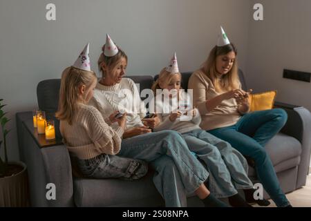 Four women and two girls, wearing party hats, sit on a sofa, using tablets and phones. A low light illuminates candles on a tray. A low-key birthday c Stock Photo