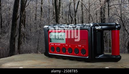 Freeze warning in effect and showing on a battery powered weather radio that has solar and hand crank backup with stiffly frozen trees behind. Stock Photo