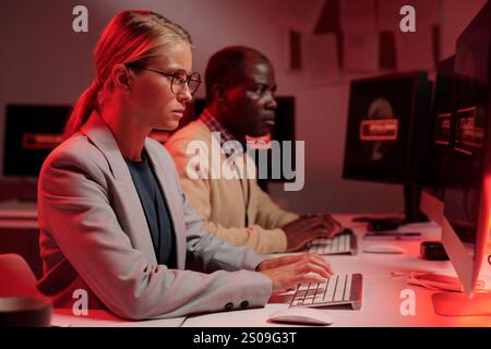 Young serious female security guard solving technical problem while sitting in front of computer screen and typing Stock Photo