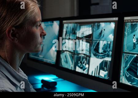 Young serious female guard sitting by workplace in front of camera and looking at computer screen with cctv videos Stock Photo