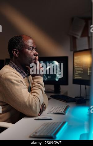 Pensive African American male programmer keeping fingers by mouth and looking at computer screen while trying to decode data Stock Photo