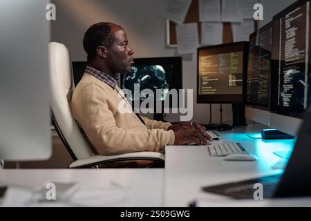 Side view of serious mature male programmer typing on computer keypad while decoding data on screen while sitting by desk Stock Photo
