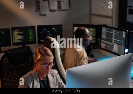 Serious young female programmer in eyeglasses and blazer looking at computer screen while decoding data against coworker Stock Photo