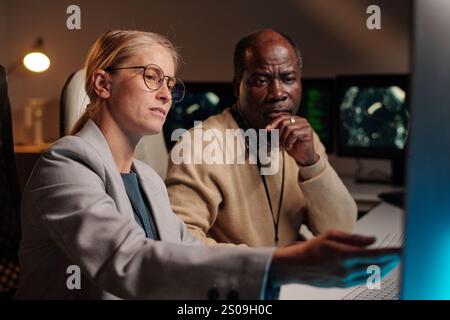 Young confident blond female IT engineer pointing at computer screen while explaining male colleague how to decode data Stock Photo