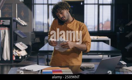 Arabian Indian male business man working busy interior designer talking with client online discuss on mobile phone fabric tissue samples write in Stock Photo