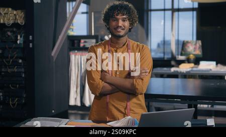 Indian Arabian man happy smiling creative designer fashion clothing tailor posing crossed hands in modern studio textile workshop with fabric color Stock Photo