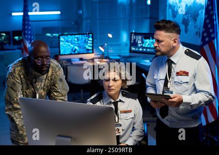 Team of serious workers of military command center looking at online data while sitting and standing in front of computer monitor Stock Photo