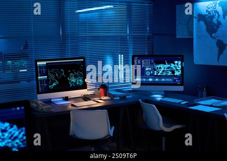 Two desktop computers with graphic data standing on desks in dark office lit with blue light in military command center Stock Photo