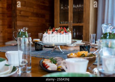 A dining table is set indoors with a large cake topped with strawberries, blueberries, and mint. Surrounding dishes include sliced meats and fruit. Stock Photo