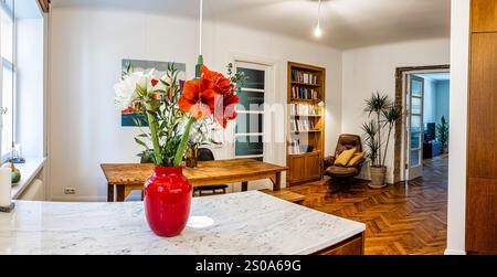 A cozy interior space features a modern kitchen with a marble countertop, vibrant red vase, wooden dining table, bookshelf, and parquet flooring. Stock Photo