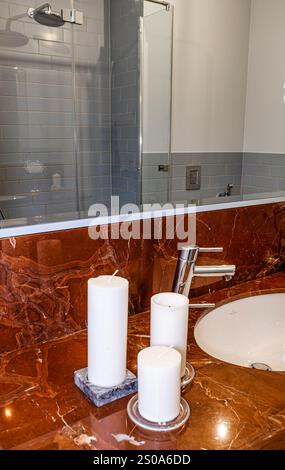 A sleek bathroom features a brown marble countertop with candles, a silver faucet, and an oval sink. A glass enclosed shower with gray tiles is visibl Stock Photo
