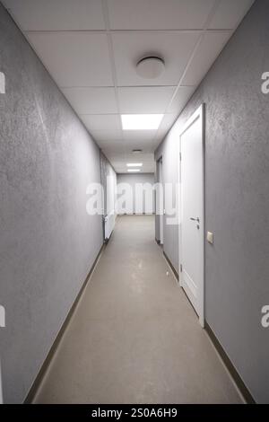 A long corridor with gray walls, light floor, and recessed lighting. Features a white door and light switch, creating a minimalist and utilitarian atm Stock Photo