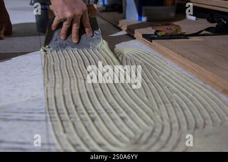 A hand uses a notched trowel to spread adhesive in parallel lines on a flat surface. Wooden planks and tools are visible, indicating a flooring projec Stock Photo