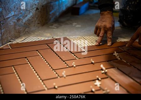 Hands position brown rectangular tiles in a herringbone pattern on a floor. White spacers ensure even spacing. Unfinished OSB walls are visible in the Stock Photo