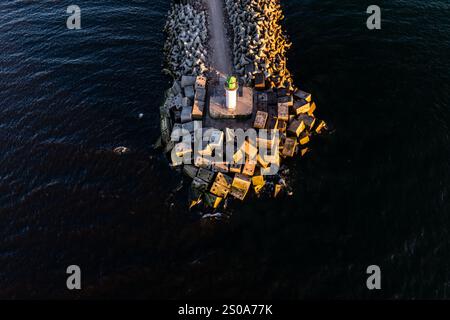 White lighthouse with green top stands on breakwater, surrounded by angular concrete blocks. Low sun angle casts long shadows over dark waters. Stock Photo