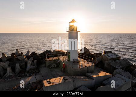 A lighthouse stands silhouetted against a setting sun on a breakwater, surrounded by concrete blocks. The ocean reflects the warm glow of the sunset. Stock Photo