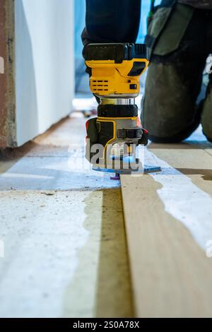 A person in dark clothing uses a yellow and black wood router on a wooden plank indoors, with visible dust and an unfinished floor in the background. Stock Photo