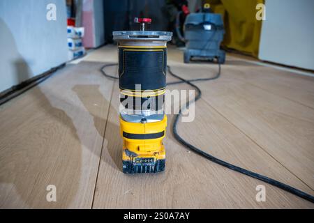 A yellow and black router sits on a wooden floor, connected by a power cord. Nearby, construction materials and tools indicate a project in progress. Stock Photo