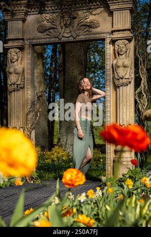 A woman stands beneath a stone archway with sculpted figures, surrounded by vibrant yellow and red tulips in full bloom, set against tall trees. Stock Photo
