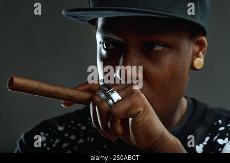 Portrait of a young man with a cool attitude smoking a cigar, wearing a baseball cap and rings, looking intensely at the camera against a dark backgro Stock Photo