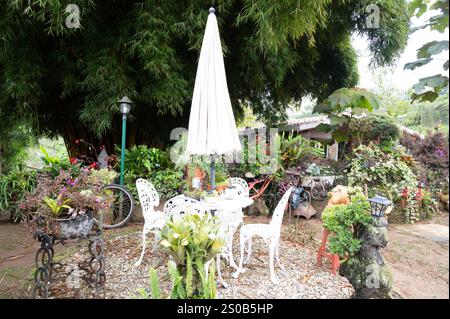 Peaceful garden oasis. White patio furniture under a parasol, surrounded by lush greenery and flowers. Stock Photo