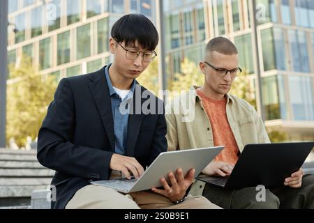 Medium shot of two male IT developers concentrated on writing code on laptops while working together outdoors in city, copy space Stock Photo