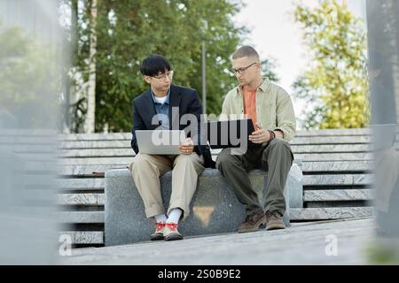 Full shot of male IT programmers using laptops during teamwork on writing code while sitting on concrete bench outside in city enjoying sunny day, copy space Stock Photo