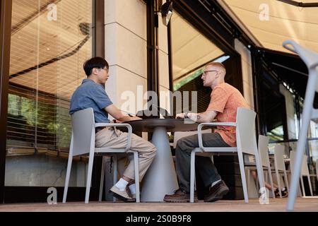 Side view of two adult male IT programmers using laptops to write code discussing work strategy while sitting at table in outdoor seating area of cafe, copy space Stock Photo