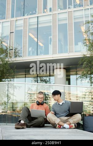 Vertical shot of diverse team of two male IT programmers working on laptops collaboratively sitting on pavement outside in city street against business building, copy space Stock Photo