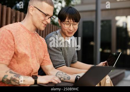 Candid shot of smiling Asian male IT programmer working on laptop collaboratively with colleague having fun while sitting on bench outside in city street, copy space Stock Photo