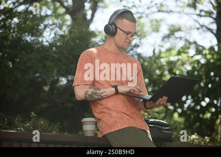 Medium shot of IT programmer in headphones using laptop to write code enjoying music and nature chilling on bench in park while freelance working outdoors, copy space Stock Photo