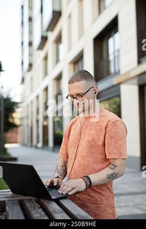 Vertical shot of smiling businessman in wire headphones participating in video conference during online work meeting while working at stand up desk outside in city street, copy space Stock Photo