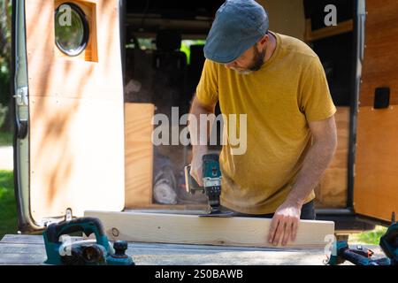 Carpenter uses cordless drill on a piece of wood near an open van, working on a woodworking project outdoors Stock Photo