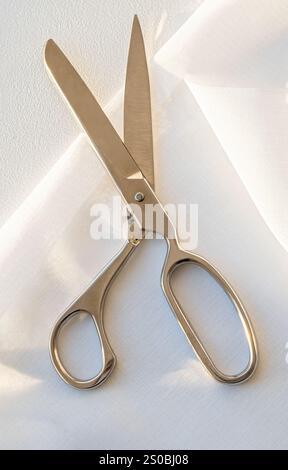 Tailor's steel scissors lying on white transparent fabric, creating contrast and highlighting the instrument's details. Close up. Vertical crop. Stock Photo