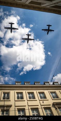 Three military aircraft flying in formation against a blue sky with fluffy clouds, viewed from below a building. Stock Photo