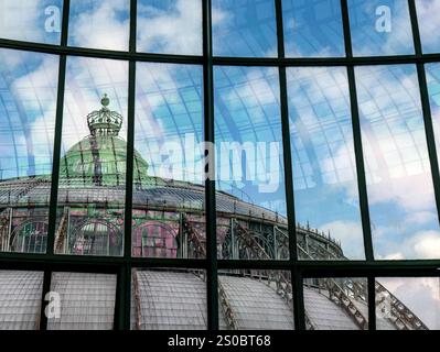 A close-up view of a glass structure reflecting a dome with a green crown, surrounded by blue sky and clouds, showcasing intricate architectural detai Stock Photo