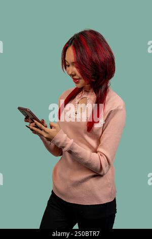 Stylish young woman with dyed red hair using smartphone, enjoying mobile technology on turquoise background Stock Photo