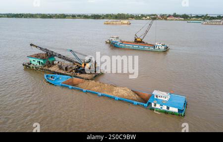 Thuong Phuoc gate, Mekong Delta, Vietnam. December 27,2024: Platforms with crane & clamshell dredge the waterway & load cargo ships/barges. Sand & gravel extraction (mining or dredging) used for concrete in construction industry raises warnings about the environmental impact of overexploitation of river bed sediments (biodiversity & ecosystem), but also about the lives of people living on riverbanks. Legal or illegal sand exploitation leads to massive erosion of banks, resulting in the loss of agricultural land but also the collapse of buildings & houses. Credit: KEVIN IZORCE/Alamy Live News Stock Photo