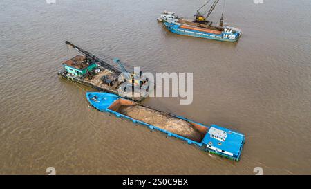 Thuong Phuoc gate, Mekong Delta, Vietnam. December 27,2024: Platforms with crane & clamshell dredge the waterway & load cargo ships/barges. Sand & gravel extraction (mining or dredging) used for concrete in construction industry raises warnings about the environmental impact of overexploitation of river bed sediments (biodiversity & ecosystem), but also about the lives of people living on riverbanks. Legal or illegal sand exploitation leads to massive erosion of banks, resulting in the loss of agricultural land but also the collapse of buildings & houses. Credit: KEVIN IZORCE/Alamy Live News Stock Photo