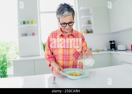Elderly woman in a colorful shirt pours milk into a bowl, creating a cozy and comfortable atmosphere at home. Stock Photo