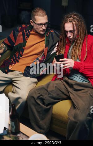 Guys sitting on sofa and playing video game on smartphone during home party Stock Photo