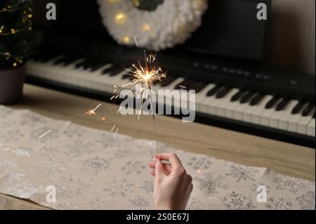 Christmas and New Year celebration. Sparkler in woman hand, piano with wreath, small tree and snowflake pattern tablecloth on the background Stock Photo