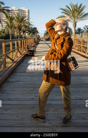 An elegant woman stands confidently on a wooden boardwalk surrounded by palm trees and modern buildings. Stock Photo