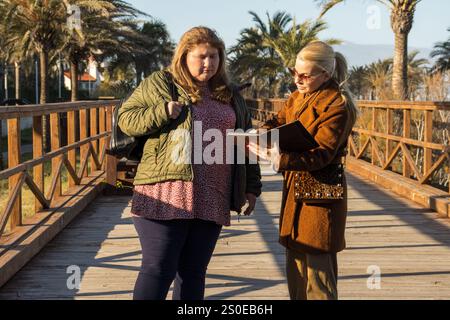 Two women stand on a wooden boardwalk, looking at something interesting displayed on a tablet. Stock Photo