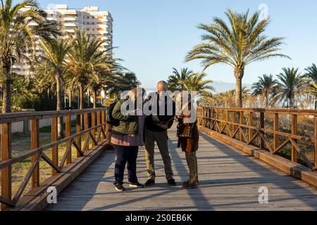 A man holds a tablet and shows information to two women while standing on a wooden boardwalk. Stock Photo