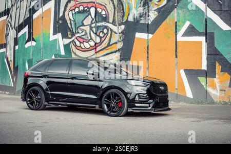 Black Ford Edge ST parked near grafiti wall. Side view of sporty SUV parked near colorful graffiti wall picturing letters and monkey. Stock Photo