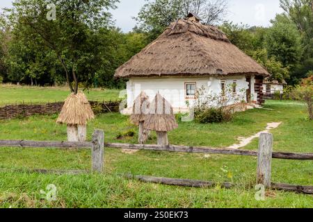 A small house with a thatched roof sits in a field. The house is surrounded by a wooden fence and a few straw bales Stock Photo