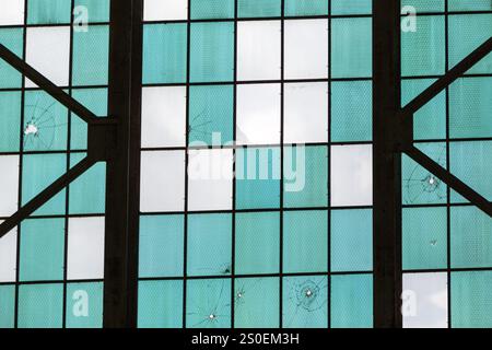 A window with a blue and white checkered pattern has been vandalized with bullet holes. The window is located in a building with a black metal frame Stock Photo