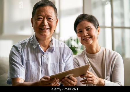 happy senior asian couple sitting on couch at home using digital tablet together Stock Photo