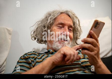 Smiling elderly man with curly gray hair, seated casually, intently using a smartphone. Stock Photo